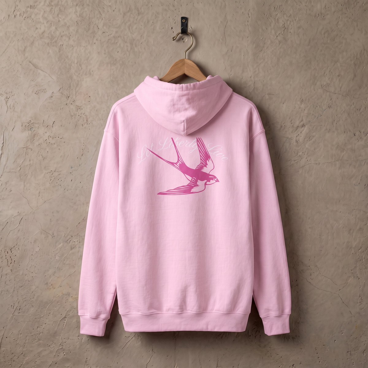 Let Liberty Live hoodie in lichtroze aan een houten hanger tegen een stucwerk muur